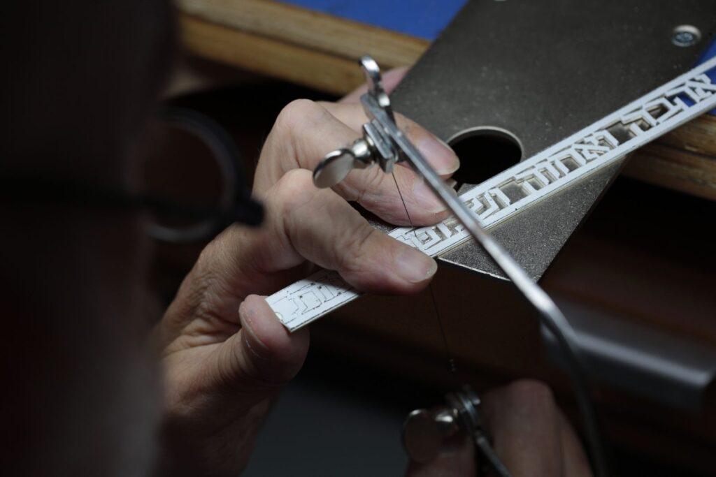 Close view of two hands guiding a jeweler's saw through a silver name pendant